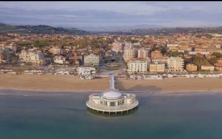 A circular pier extending into the water with a building on top, connected to the mainland by a walkway. The background shows a beach and a cityscape with various buildings.