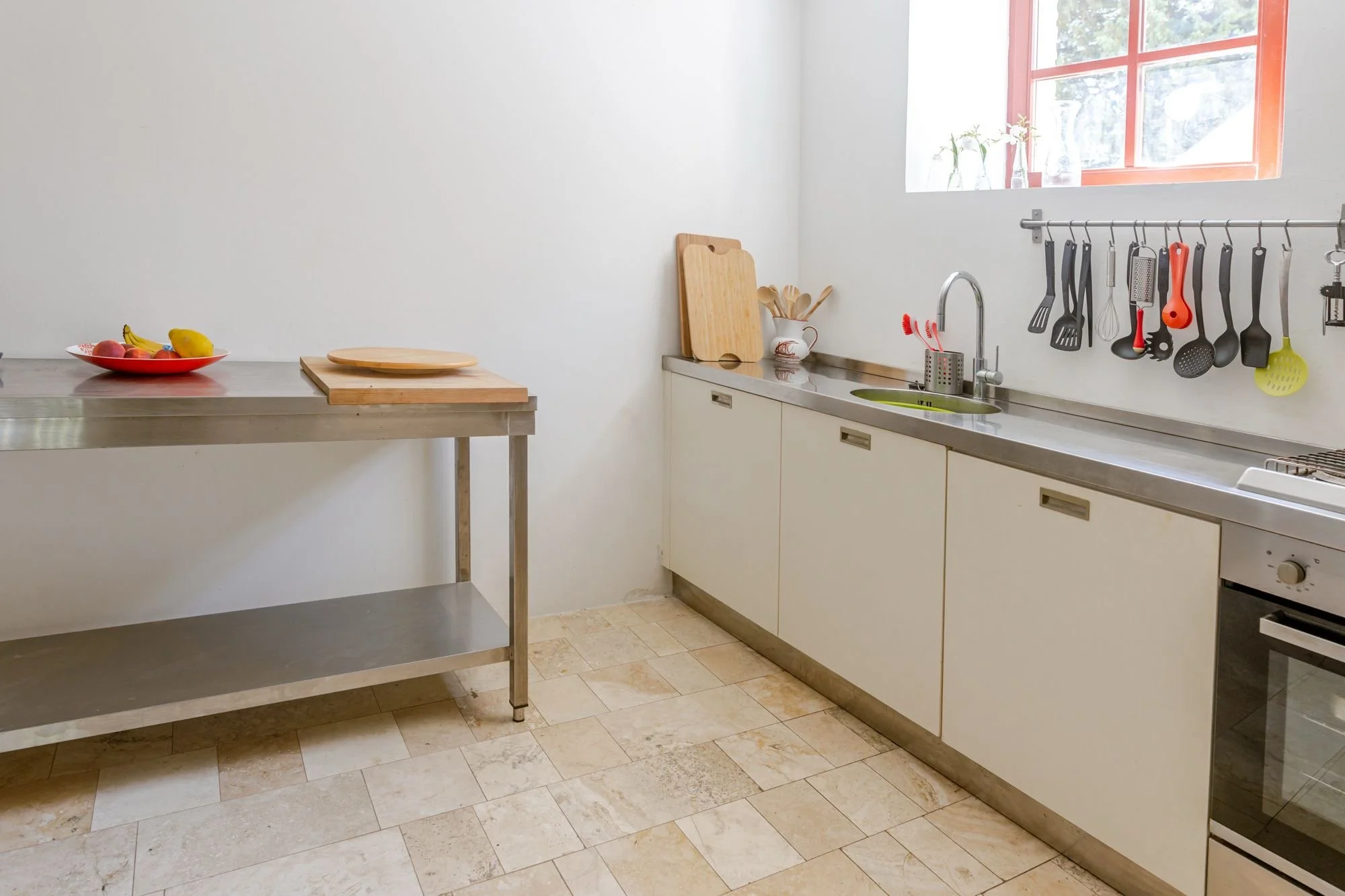 A kitchen with white cabinets, a stainless steel countertop, a window with orange trim, and a variety of kitchen utensils hanging on a rail. There is a bowl of fruit on a separate stainless steel table.