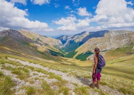Hiker standing on a trail in a mountainous landscape with green hills, rocky peaks, and a blue sky with clouds.