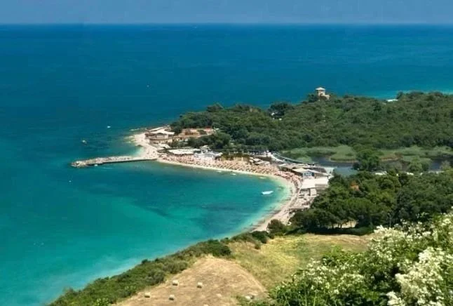 Aerial view of a coastal area with a beach, turquoise waters, and greenery, including trees and hills, near a small town or resort.