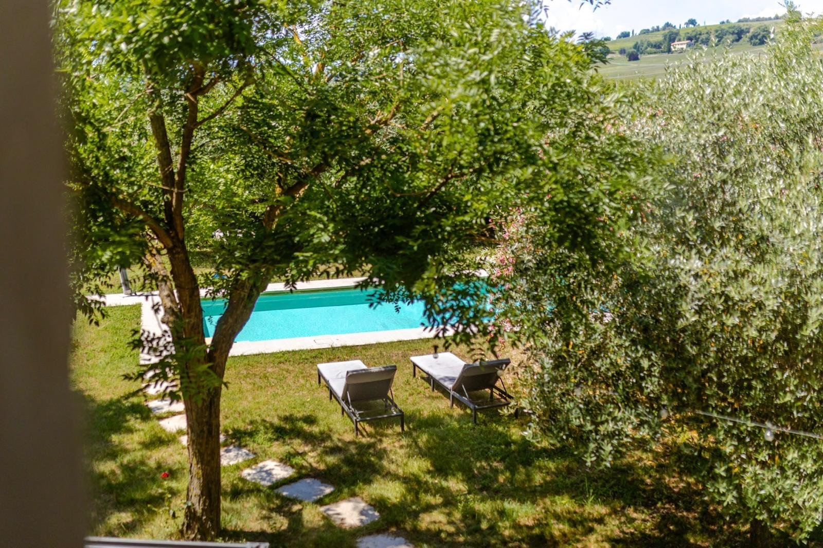 View of a backyard with a swimming pool, two lounge chairs, and trees providing shade, with hills in the background.