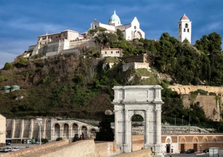 View of a historic hilltop city with a white church and bell tower, a stone arch, and Mediterranean-style buildings under a blue sky.