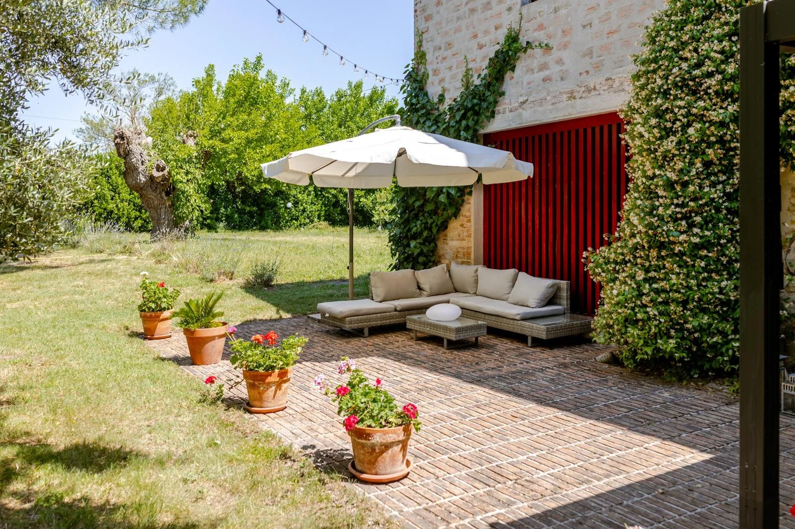 Outdoor patio with wicker sectional sofa with beige cushions, white umbrella, potted plants, brick flooring, lush green trees, and a vine-covered brick wall.
