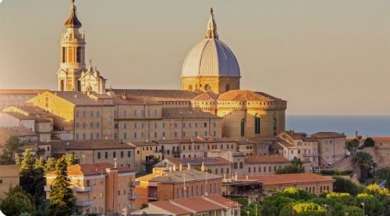 Sunset view of historical European city with domed cathedral and bell tower, surrounded by orange-roofed buildings.
