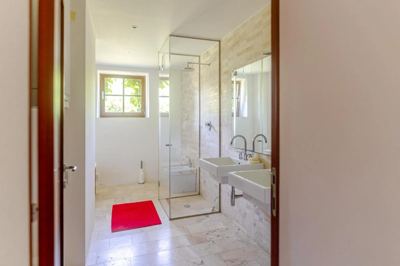 Bright bathroom with window, glass shower enclosure, wall-mounted sinks, mirror, red rug, and beige tile flooring.