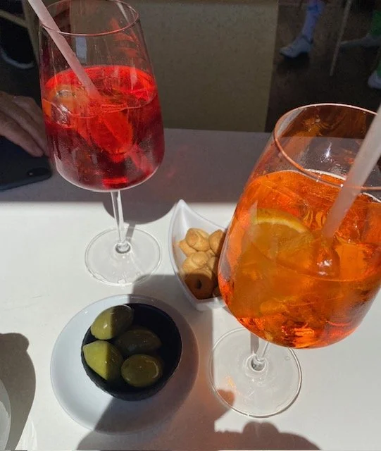Two tall glasses of orange and pink beverages with ice and straws on a white table, accompanied by small bowls of green olives and peanuts.