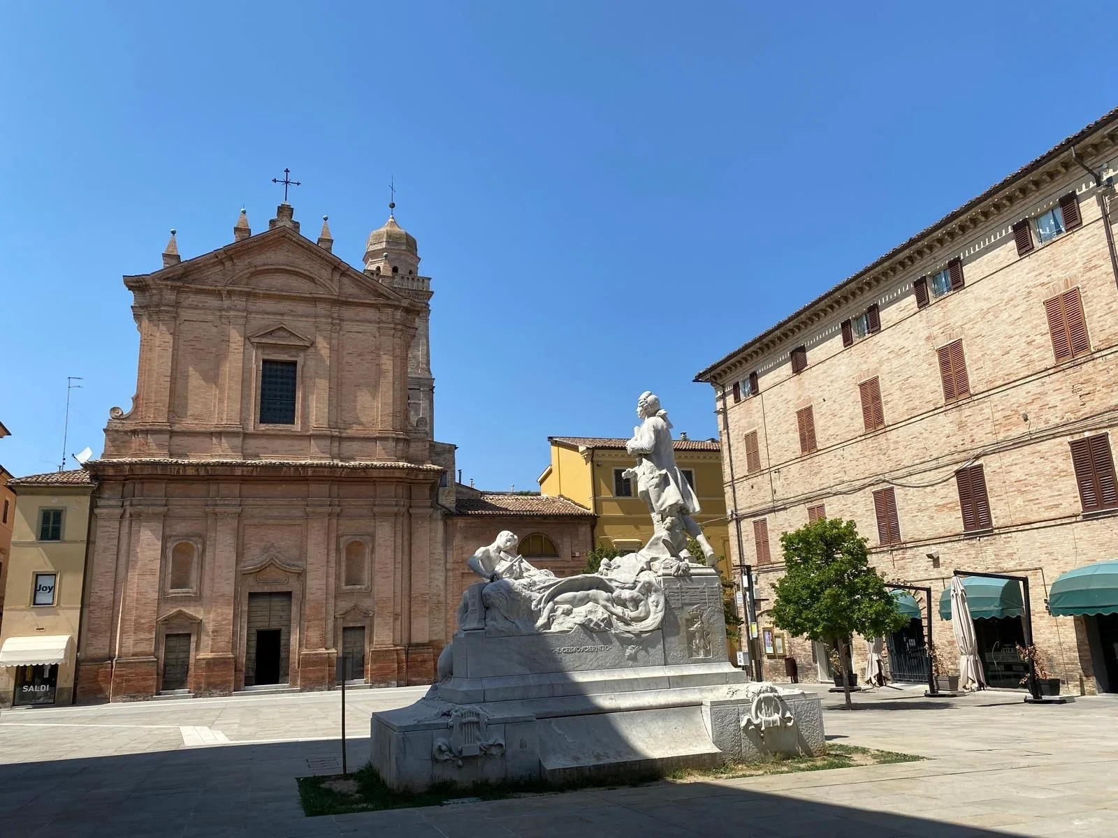 A marble statue in a public square with a church in the background, under a bright blue sky.