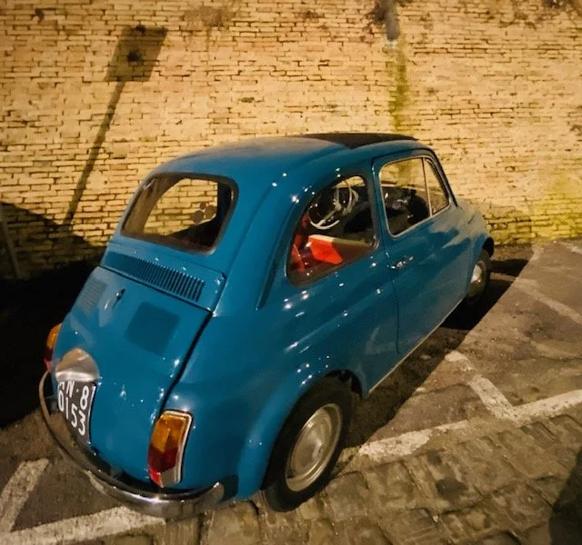 Blue vintage microcar parked in a parking lot at night against a brick wall.