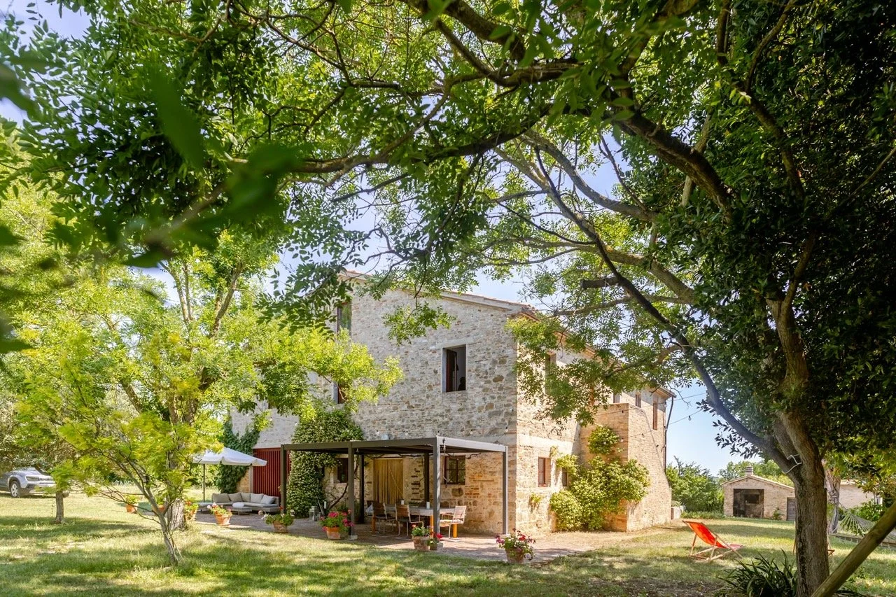 Stone house with a patio, surrounded by trees and greenery on a sunny day.