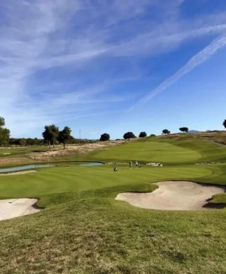 A golf course with green fairways, sand bunkers, a small pond, and several golfers playing under a partly cloudy blue sky.