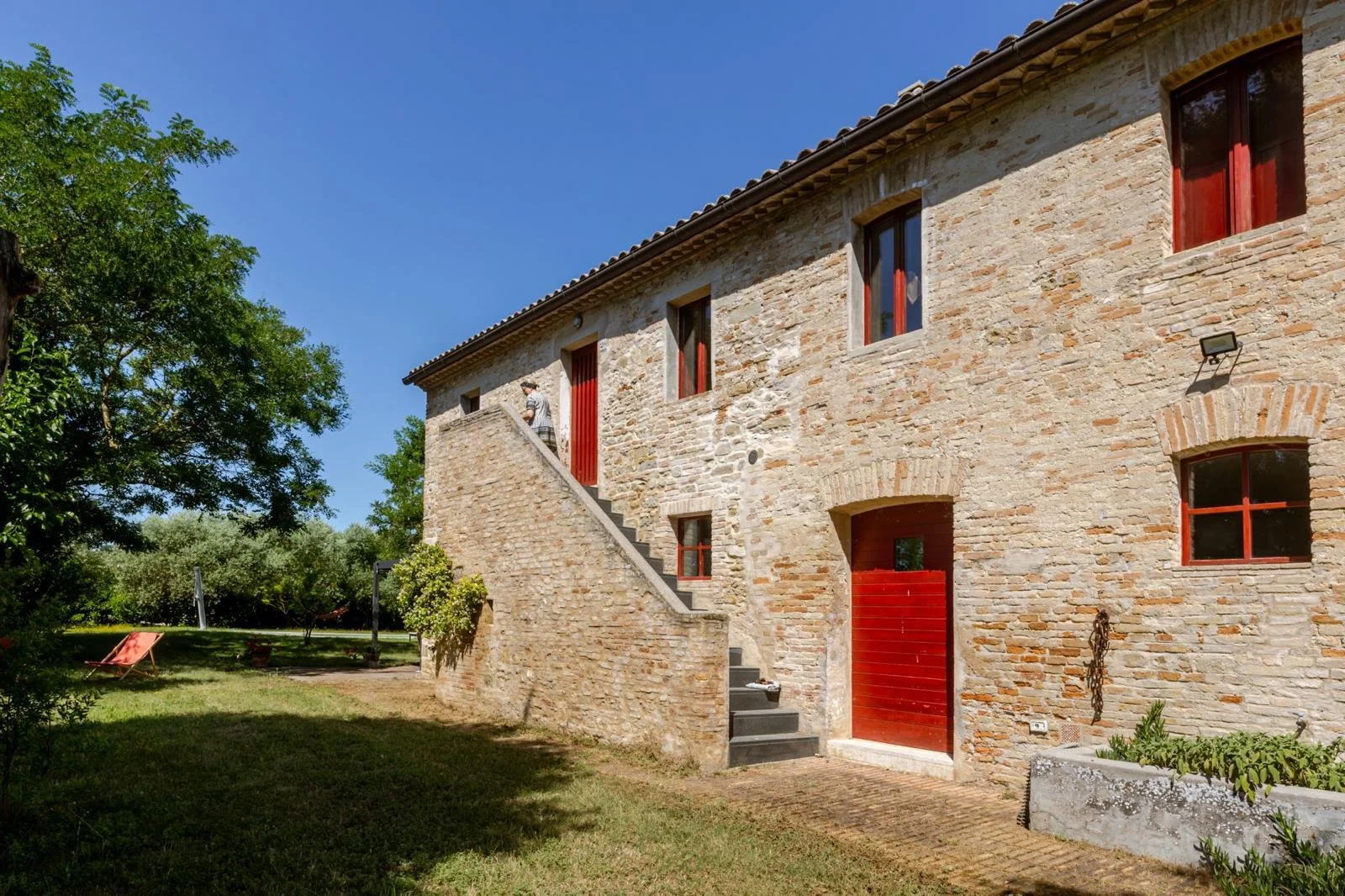 A stone house with red window frames and a red door, exterior staircase leading to a second-floor entrance, surrounded by greenery, under a clear blue sky.