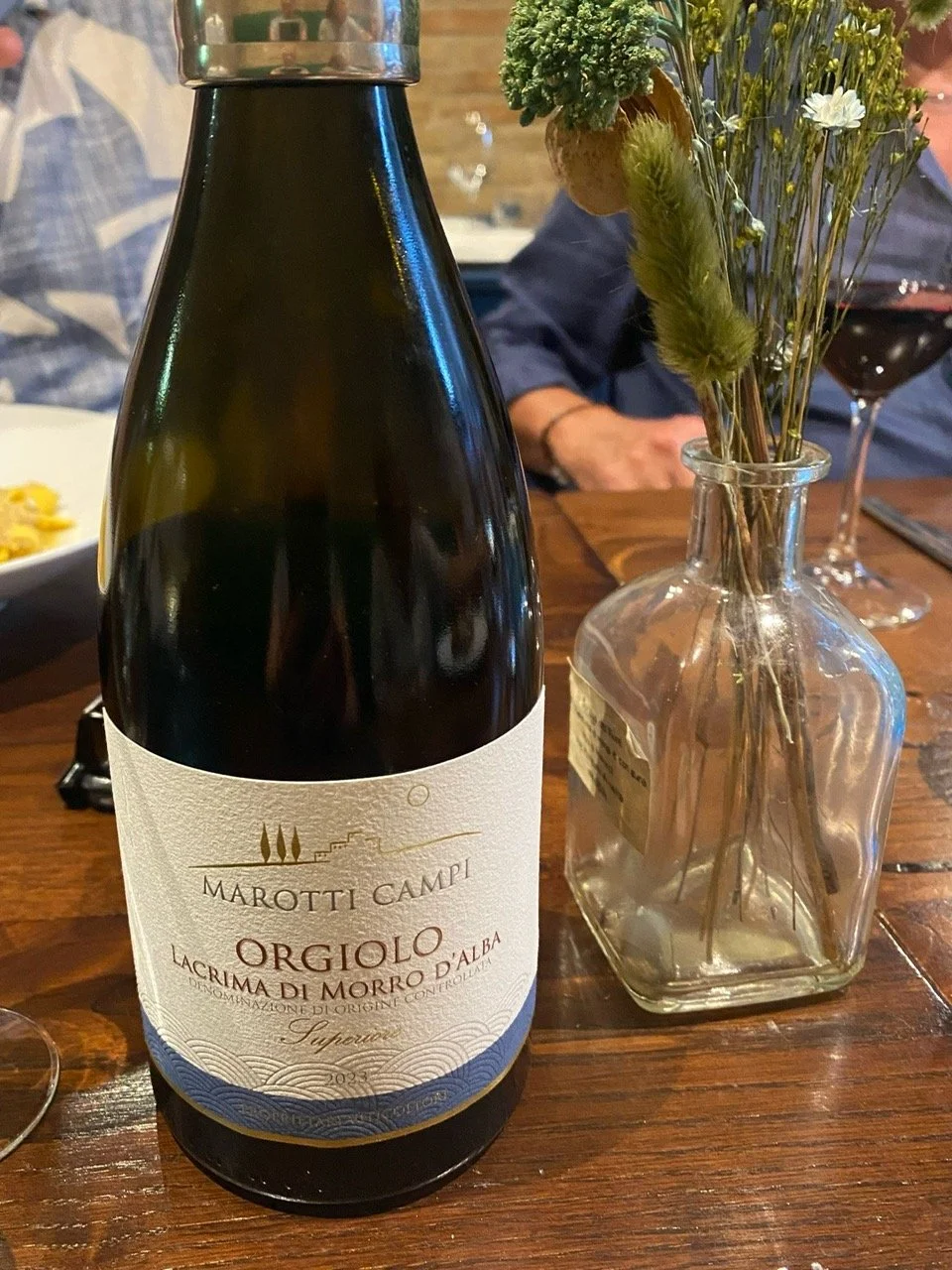 A bottle of Marotti Campi Orgiolo Lacrima di Morro d'Alba wine on a wooden table next to a glass of red wine and a clear glass vase with mixed wildflowers.