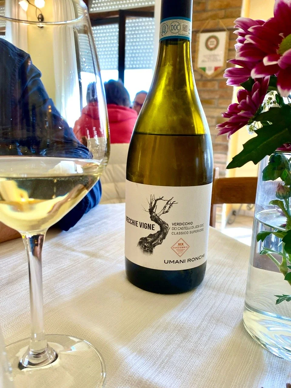 A bottle of white wine, a glass of white wine, and a vase with pink and purple flowers on a dining table in a restaurant.