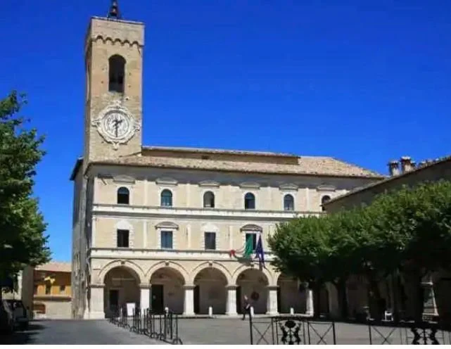 A historic church with a tall clock tower, arched ground level entrance, and a clear blue sky in the background. Trees and a paved area with fencing are in the foreground.