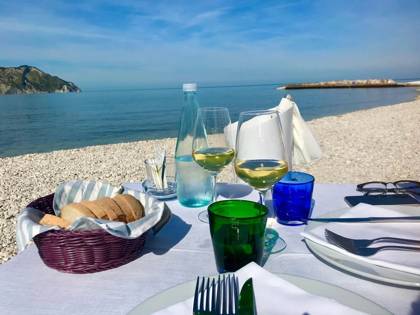 A table set for a meal on a beach, with glasses of white wine, a basket of sliced bread, water bottles, cutlery, and colorful glasses, overlooking the ocean with a distant island and a breakwater in the background.