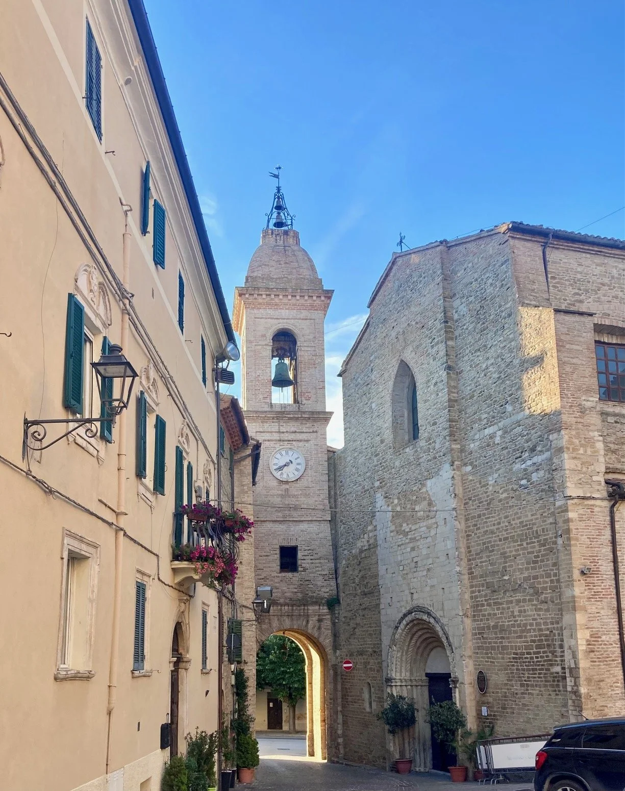 A narrow street in a European town with historic stone and stucco buildings, a clock tower, and an archway at the end of the street under a clear blue sky.