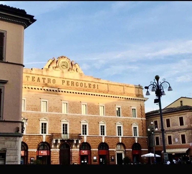 The facade of Teatro Pergolesi, a historic theater in Italy, with red and white brick exterior, large arched windows on the ground floor, and decorative architectural details under a clear blue sky.