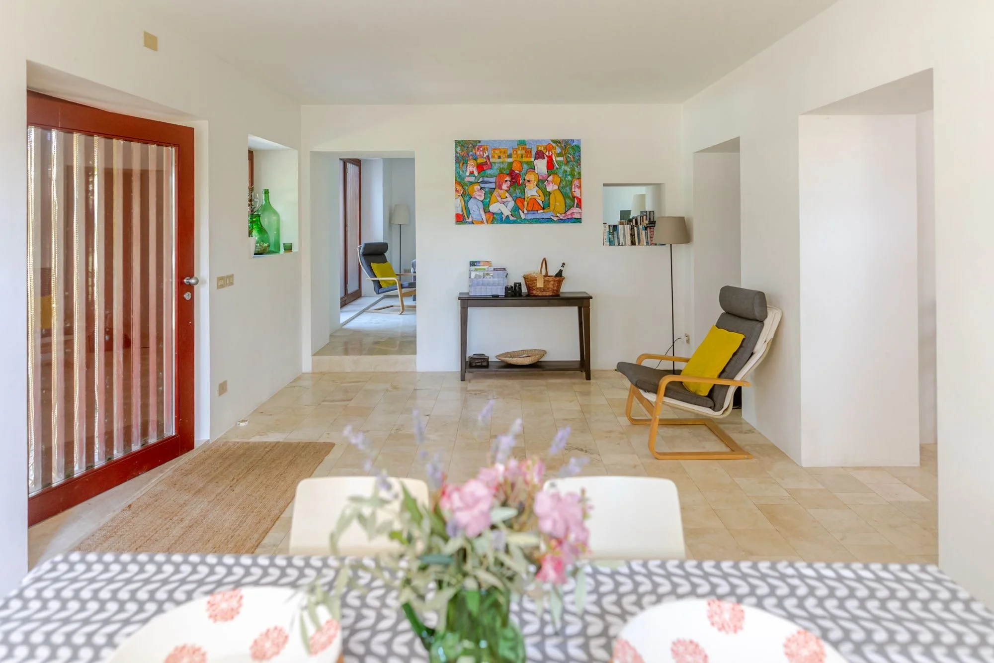 Cozy living room with a white couch, armchair with yellow pillow, painting on wall, wooden console table, bookshelf, and floor lamp. Front door with glass panel, and view into an additional room with a chair and window.