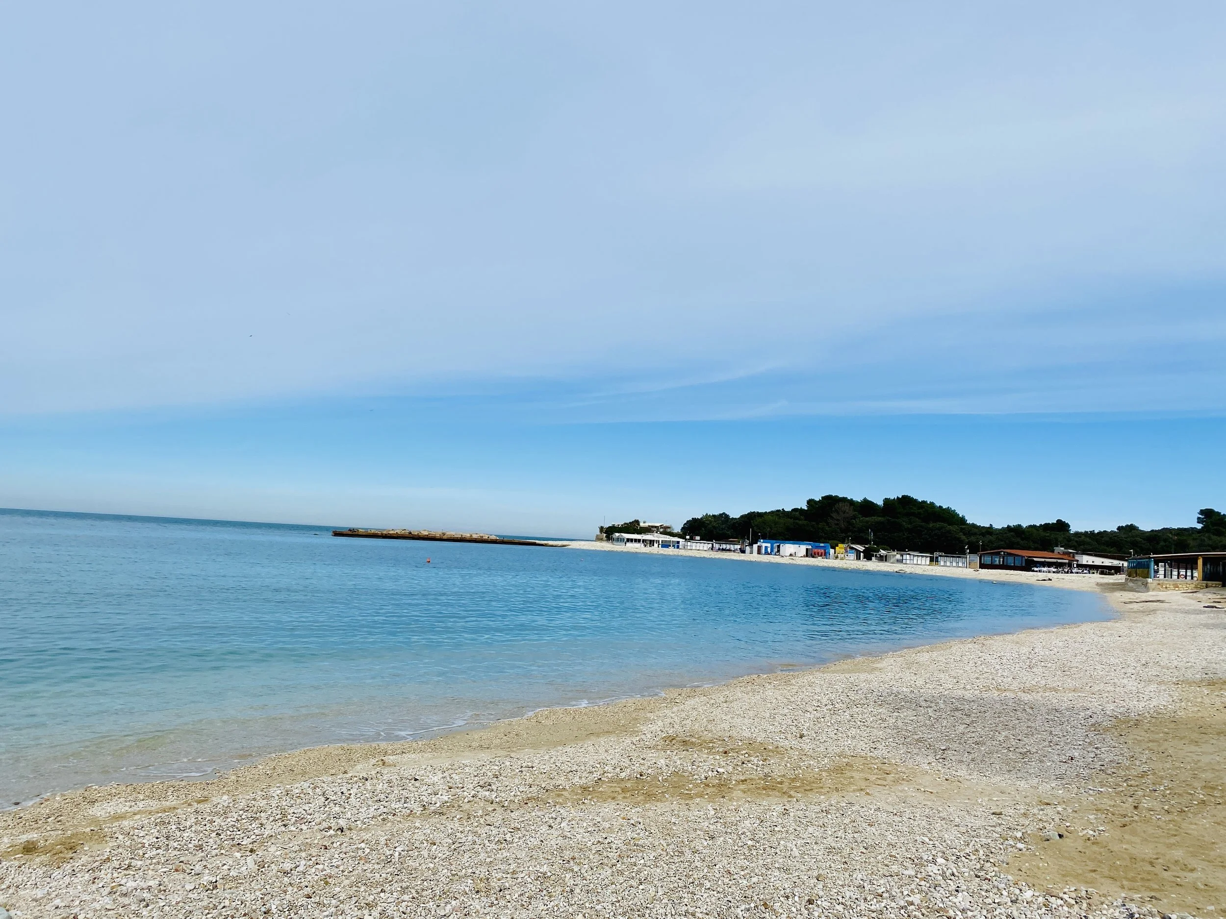 A tranquil beach scene with pebbled shoreline, clear blue water, and a distant pier with small buildings and trees along the coast under a partly cloudy sky.