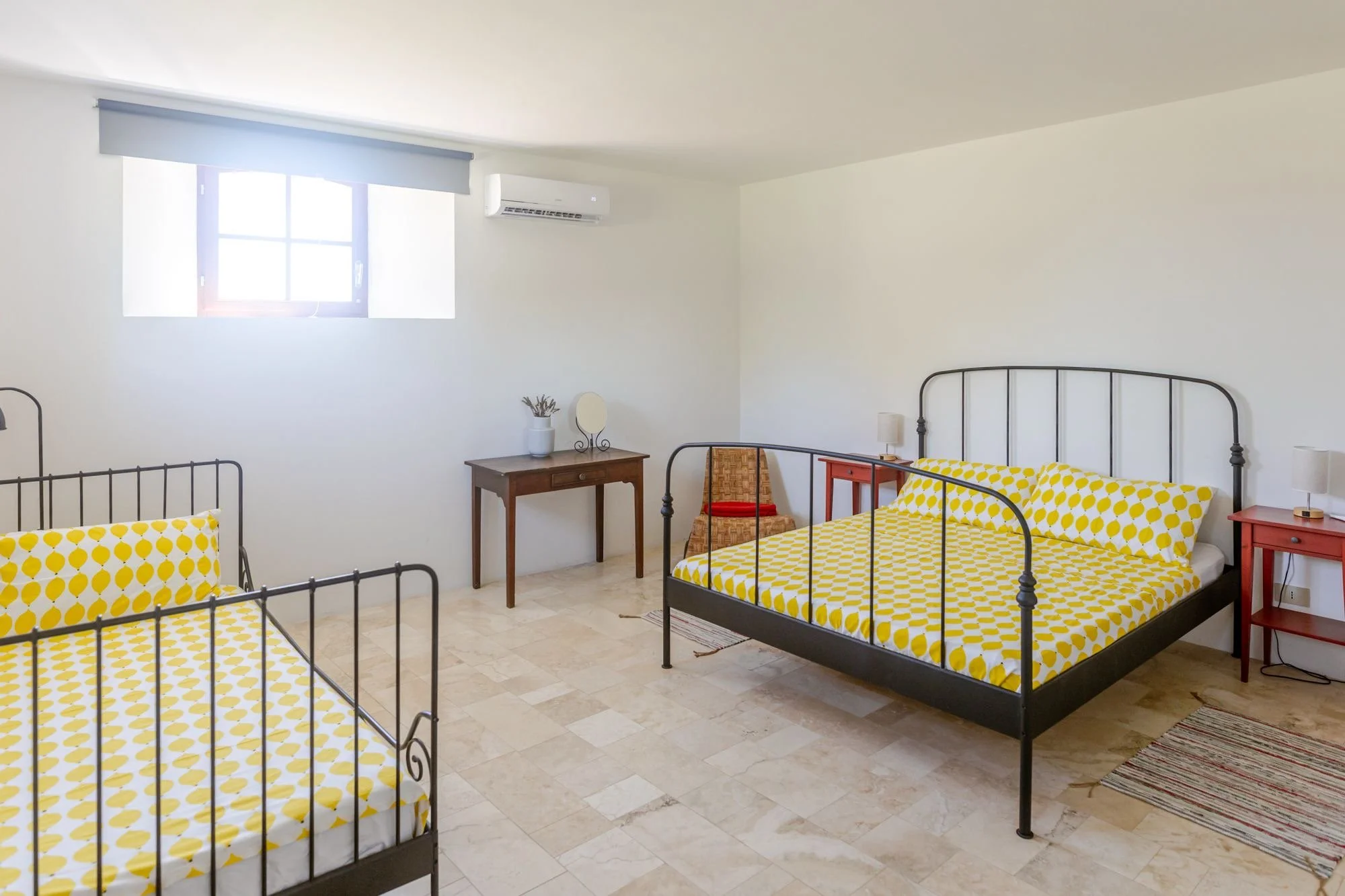 A bright room with two black metal twin beds with yellow polka dot bedding. A small wooden table with a white vase and a mirror, a red side table, and a wicker chair. A window with a blue valance and an air conditioning unit. Light-colored tiled floor and a woven rug.