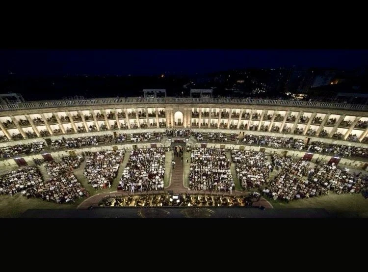 Nighttime view of an outdoor amphitheater filled with an audience seated in rows, surrounded by a multi-story building with lit balconies.