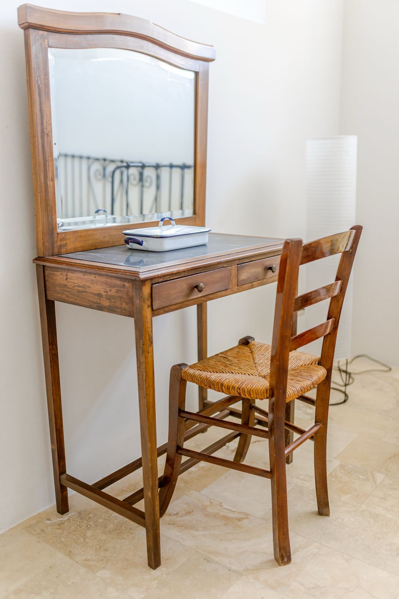 A wooden dressing table with a mirror attached, along with a wooden chair with a woven seat, positioned against a light-colored wall. There is also a small, rectangular, white container with a metal handle on the table.