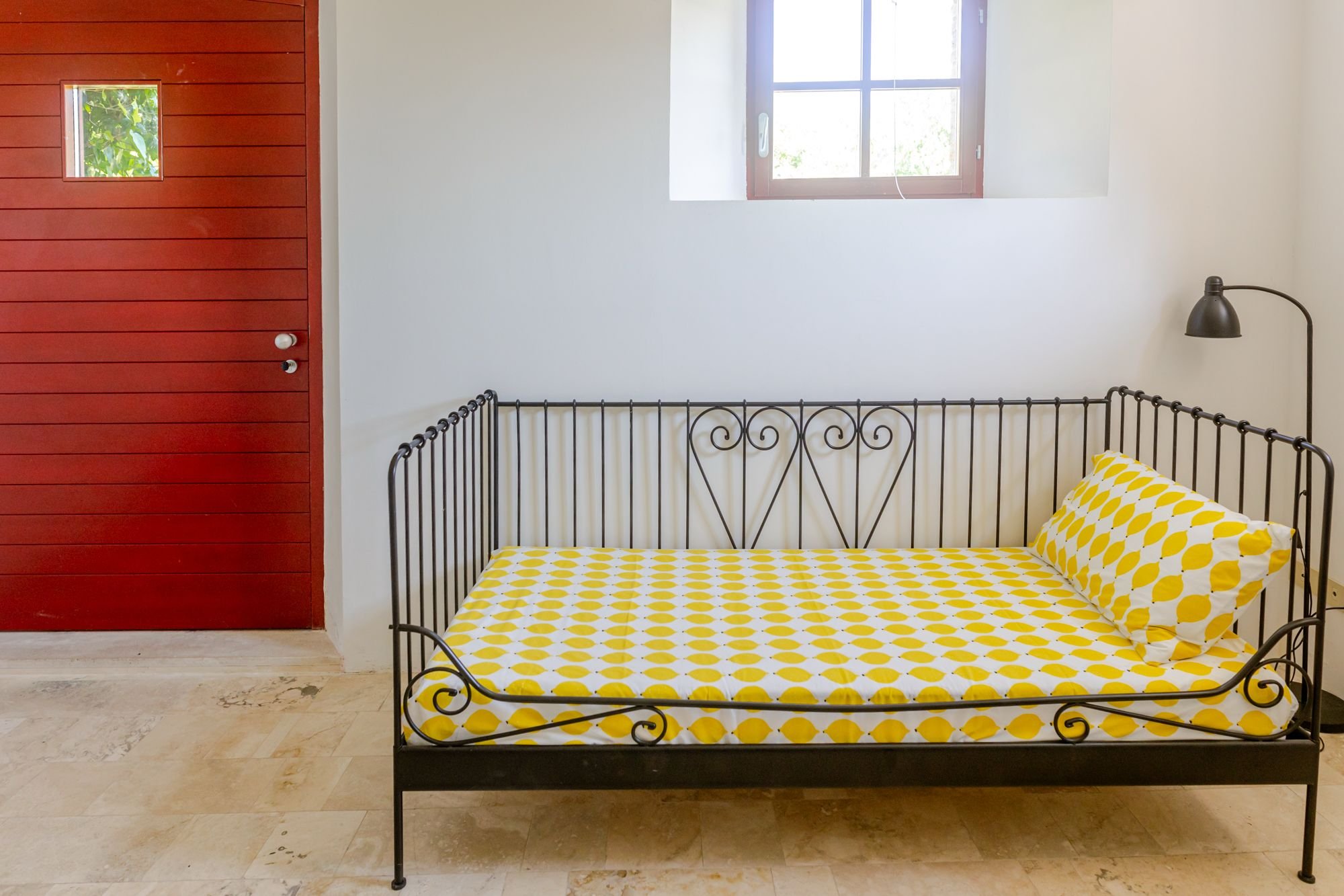 A bedroom featuring a black metal bed frame with yellow and white polka dot bedding, a matching pillow, a wall-mounted black reading lamp, a window with a wooden frame, and a red wooden door with small windows.