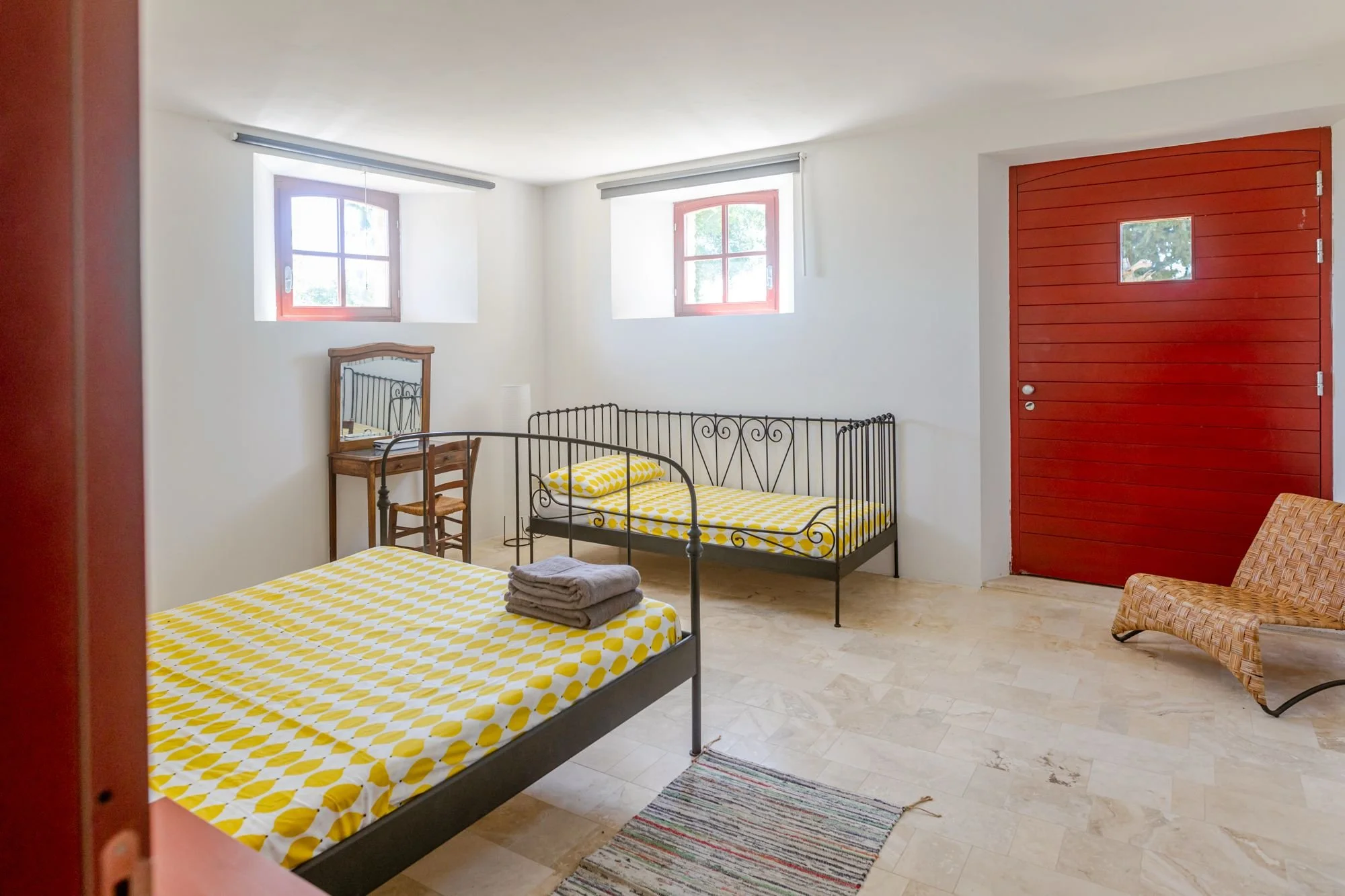 A bedroom with two metal-framed beds with yellow and white polka dot bedding, a small wooden vanity with a mirror, a wicker chair, and a red wooden door. There are two windows letting in natural light.