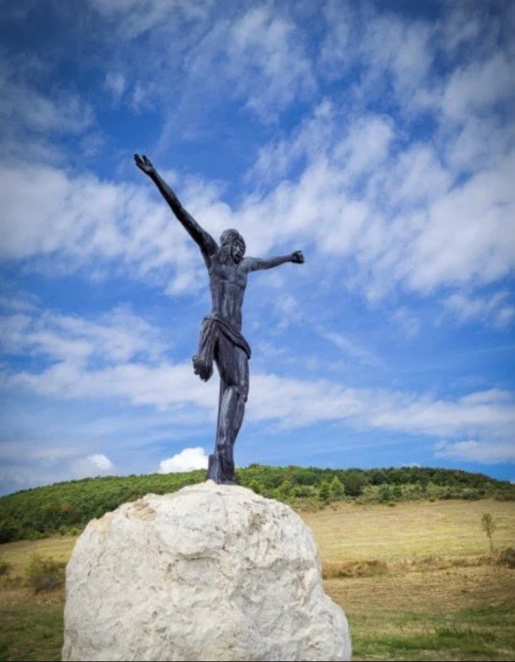 Statue of Jesus Christ with arms outstretched on a rock, in a grassy landscape under a partly cloudy blue sky.