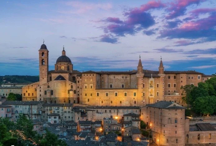 Historic stone castle with towers and domes at sunset, surrounded by smaller buildings with lights, and green trees nearby.