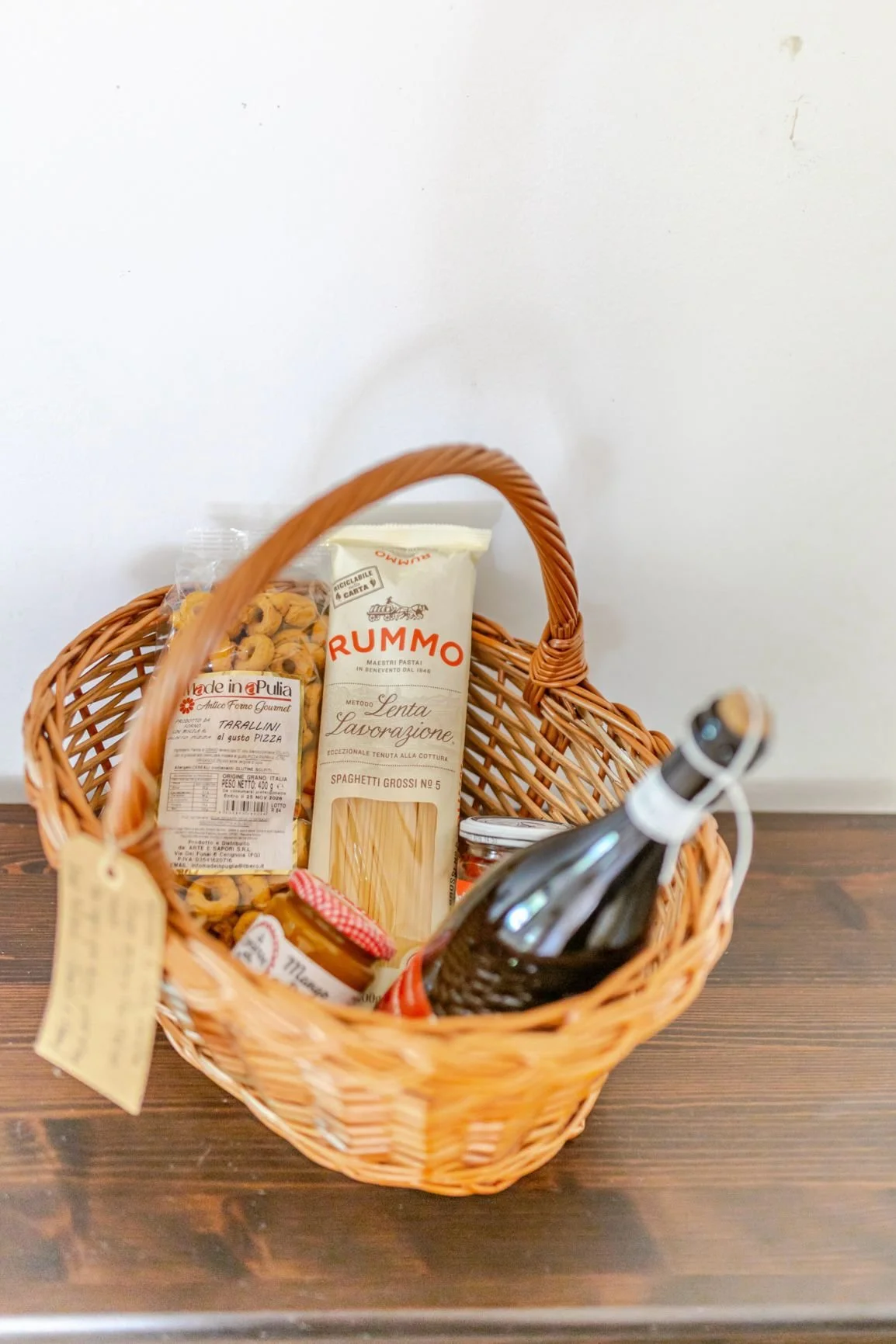 Wicker basket containing a bag of pasta, a jar of sauce, and a bottle of wine placed on a wooden surface against a white wall.