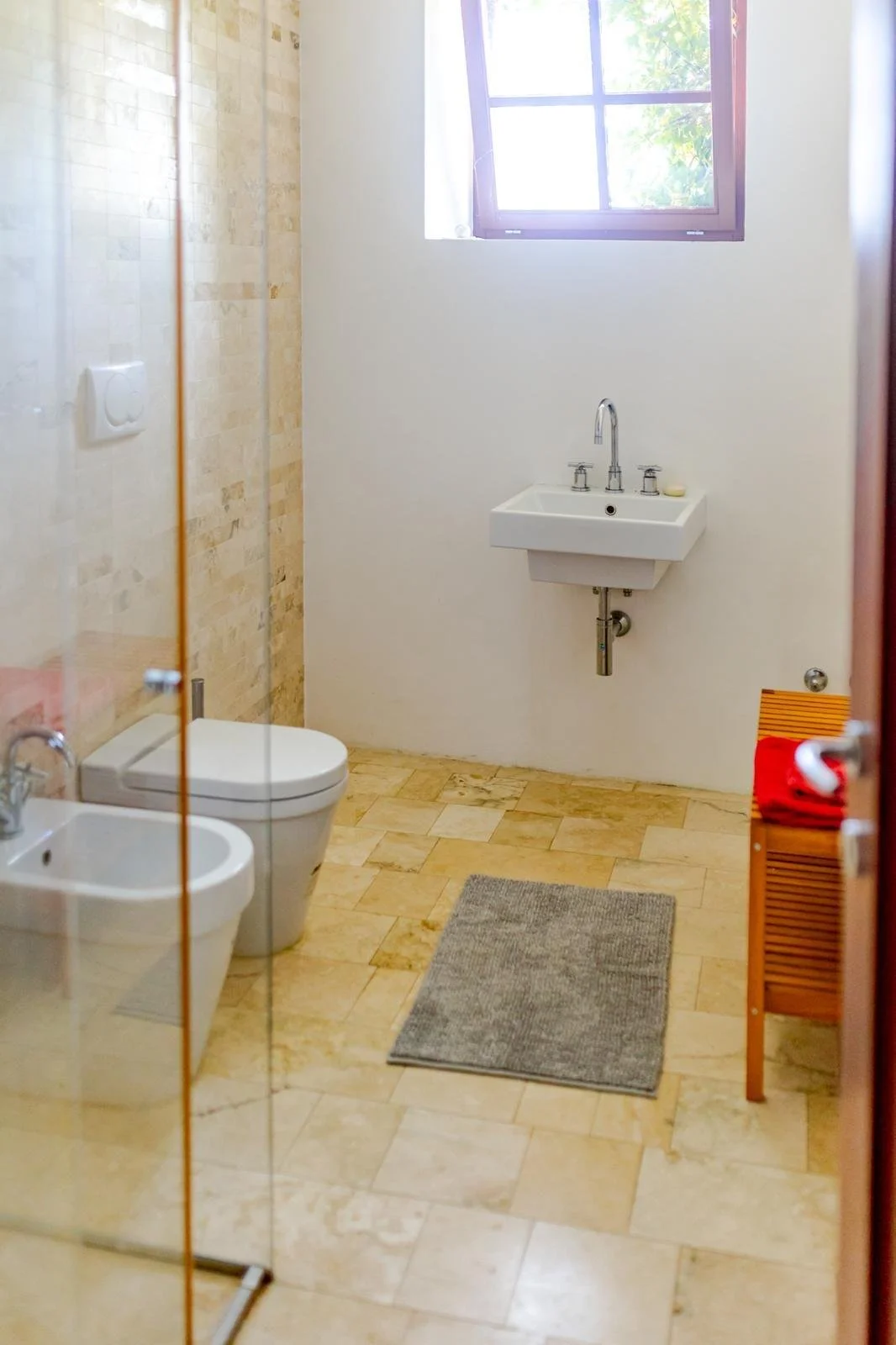 First look at a minimalist bathroom with a small sink, toilet, bidet, and a wooden bench with red towels, a window with natural light, and beige tiled floor and wall, with a gray bath mat.