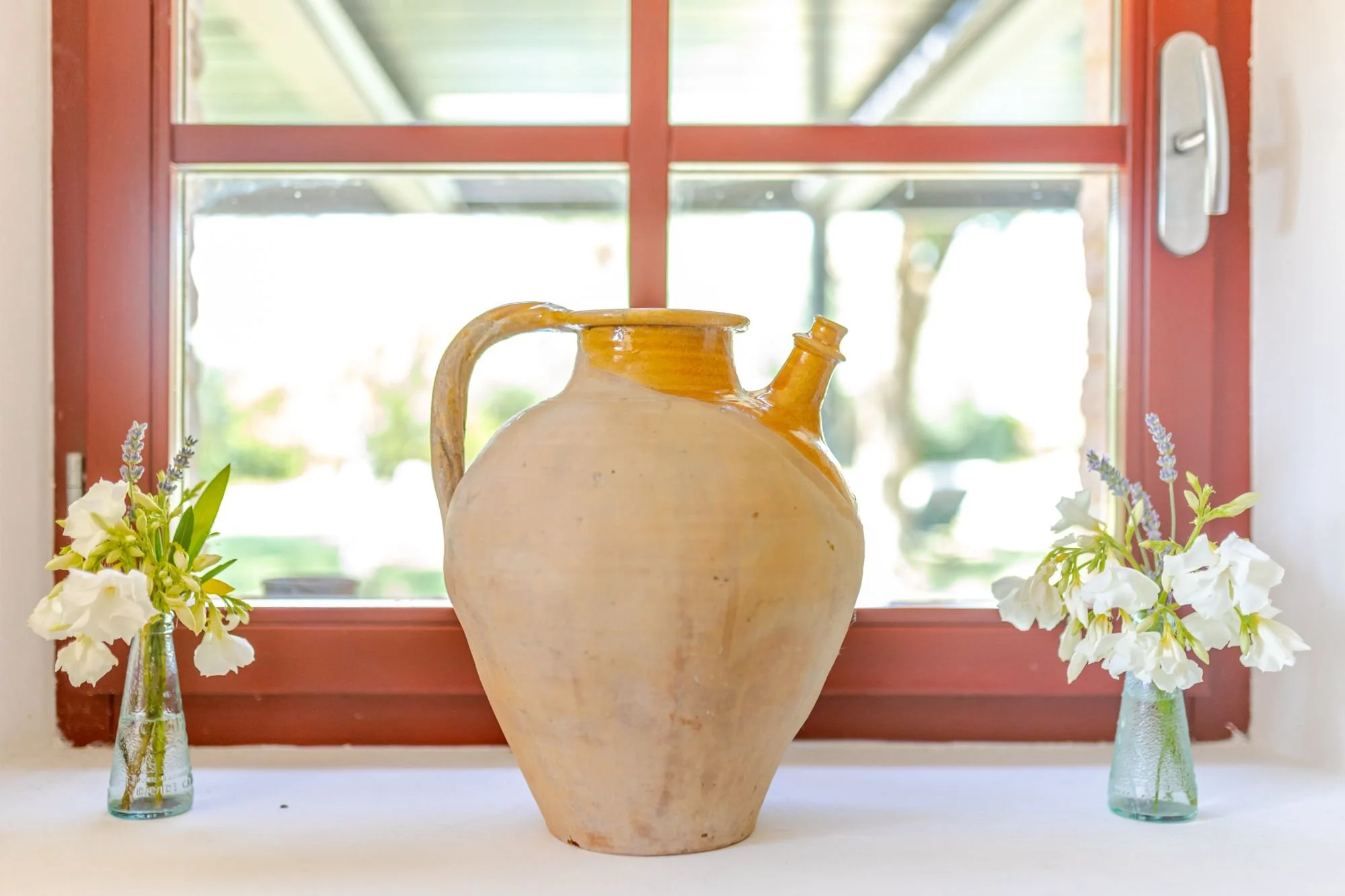 A large ceramic vase with a handle, placed on a window sill with white flowers in glass vases on either side. A window with red framing and a view of trees outside is in the background.