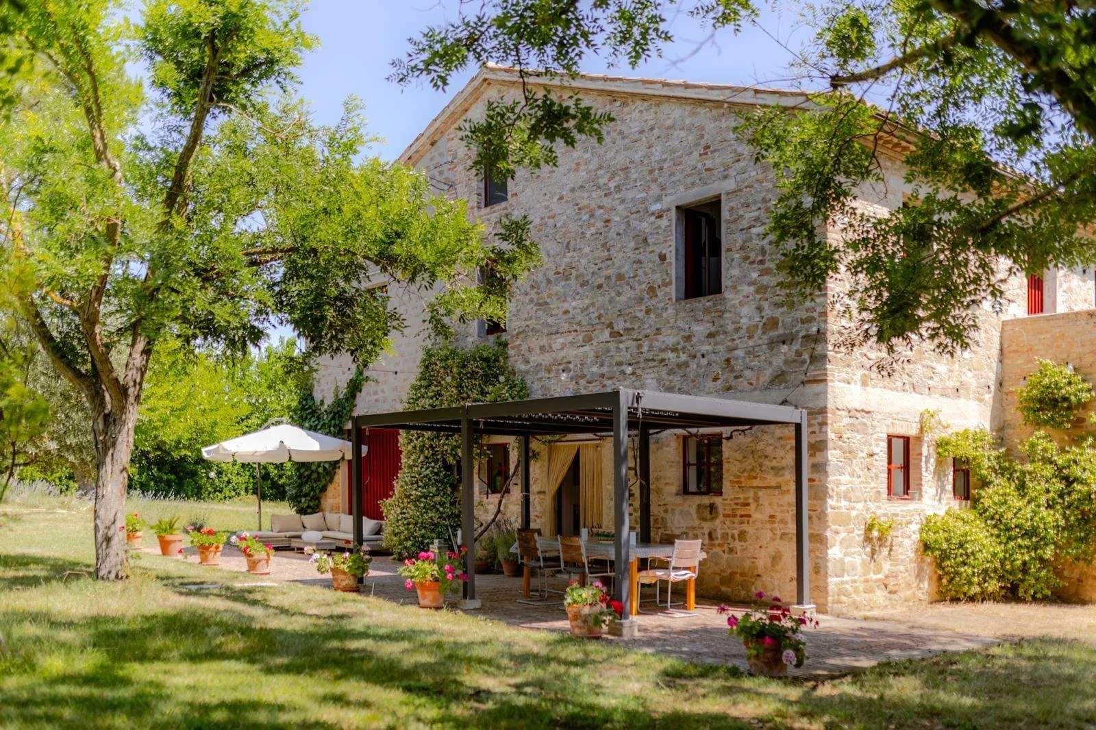 A stone house with multiple windows and a patio area with outdoor furniture, potted plants, and an umbrella, surrounded by greenery and trees under a blue sky.