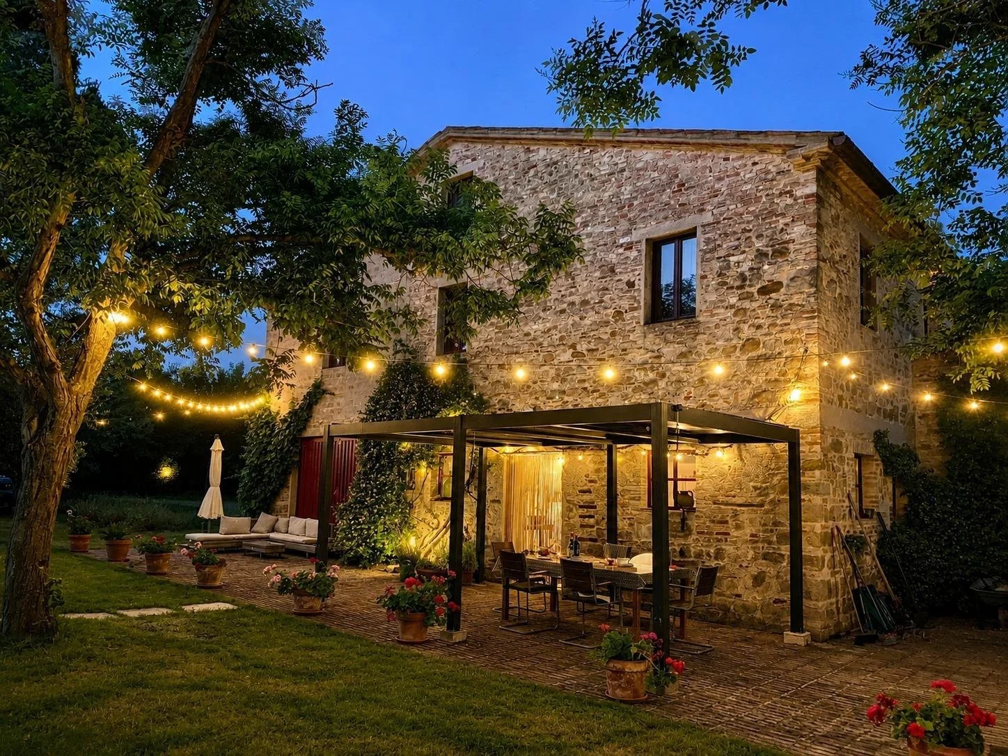 An outdoor patio area at night with string lights hanging above, a covered dining table with chairs, potted plants, a stone house with illuminated windows, and a grassy lawn with trees.