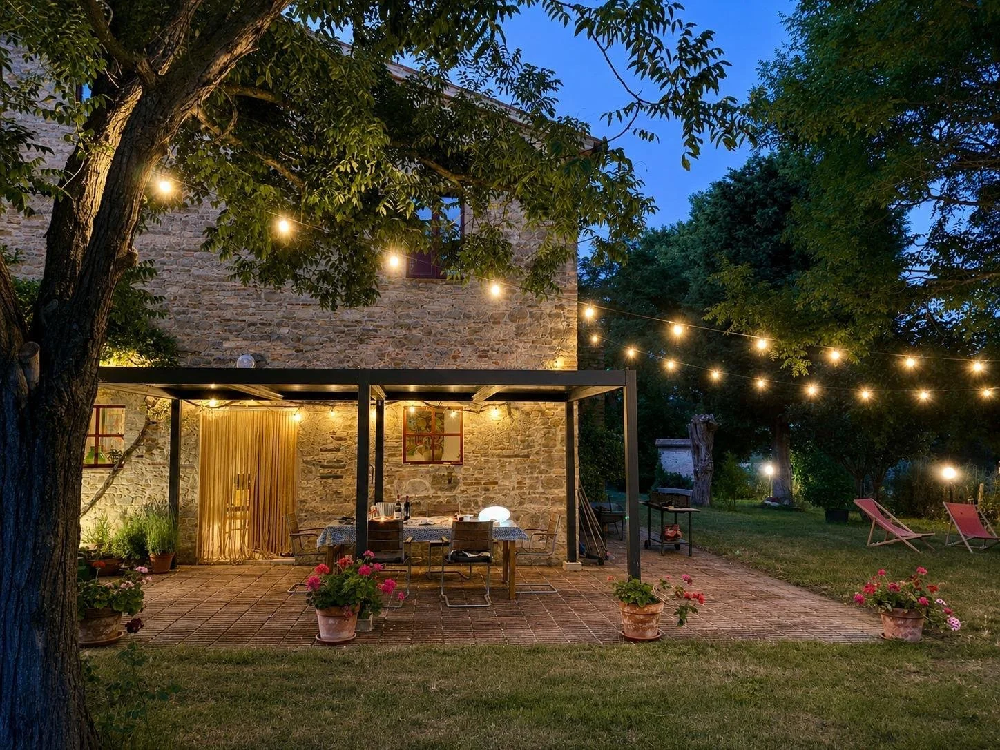 A cozy outdoor patio area at dusk with string lights overhead, a stone house in the background, a table set with a tablecloth and chairs, potted plants, and lawn chairs on the grass.