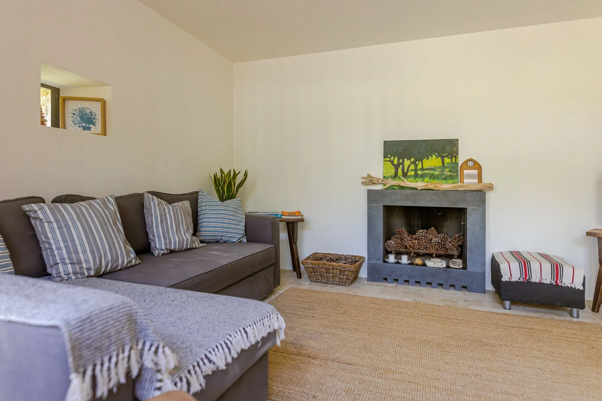 Living room with gray sofa, striped pillows, potted plant, fireplace with artwork and small decorative items, woven basket, and striped bench, decorated in neutral tones with a focus on natural materials.
