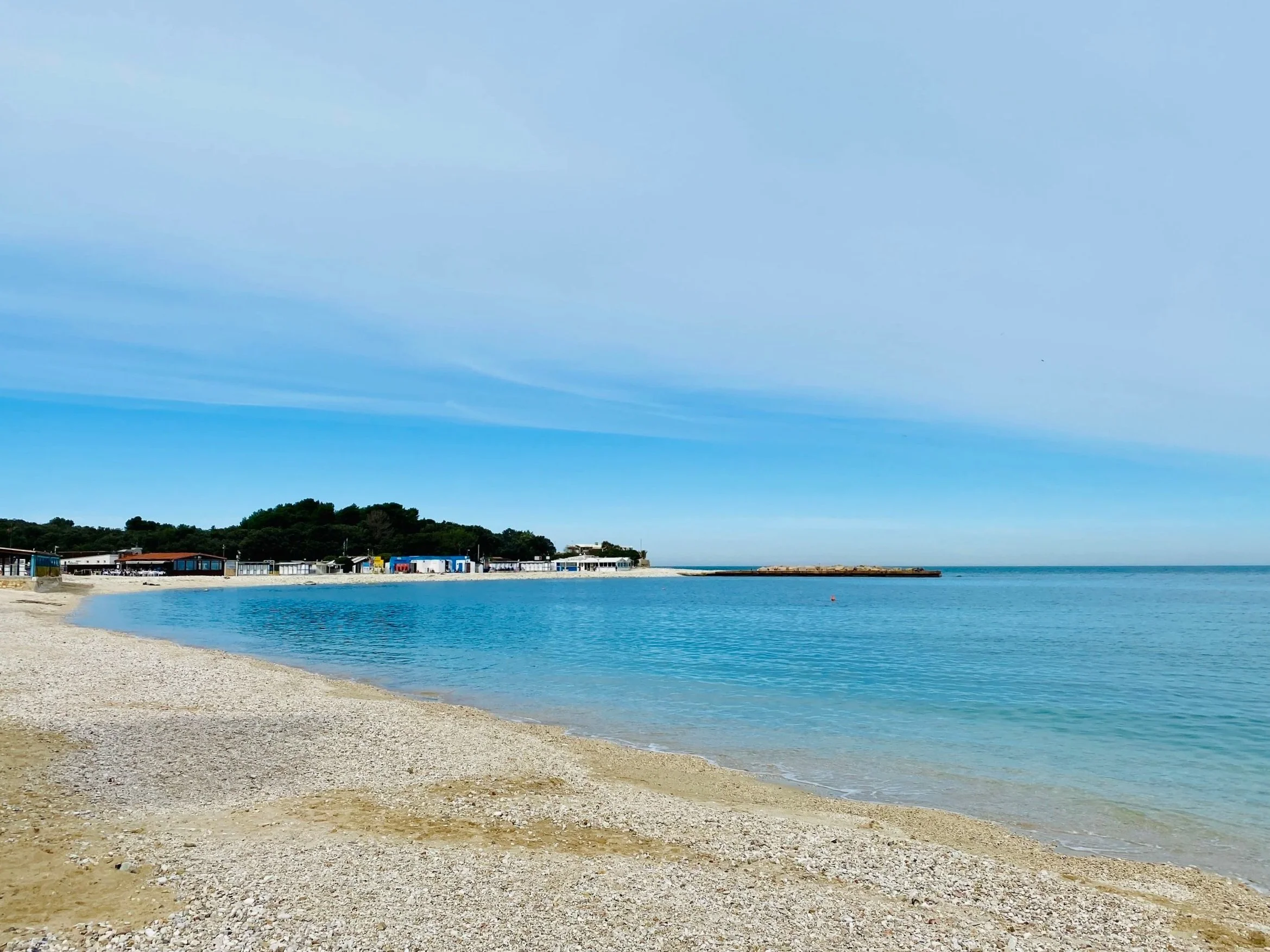 A peaceful beach scene with a sandy shoreline, calm blue water, and a clear sky with light clouds. In the background, there are some buildings and greenery on a hill.