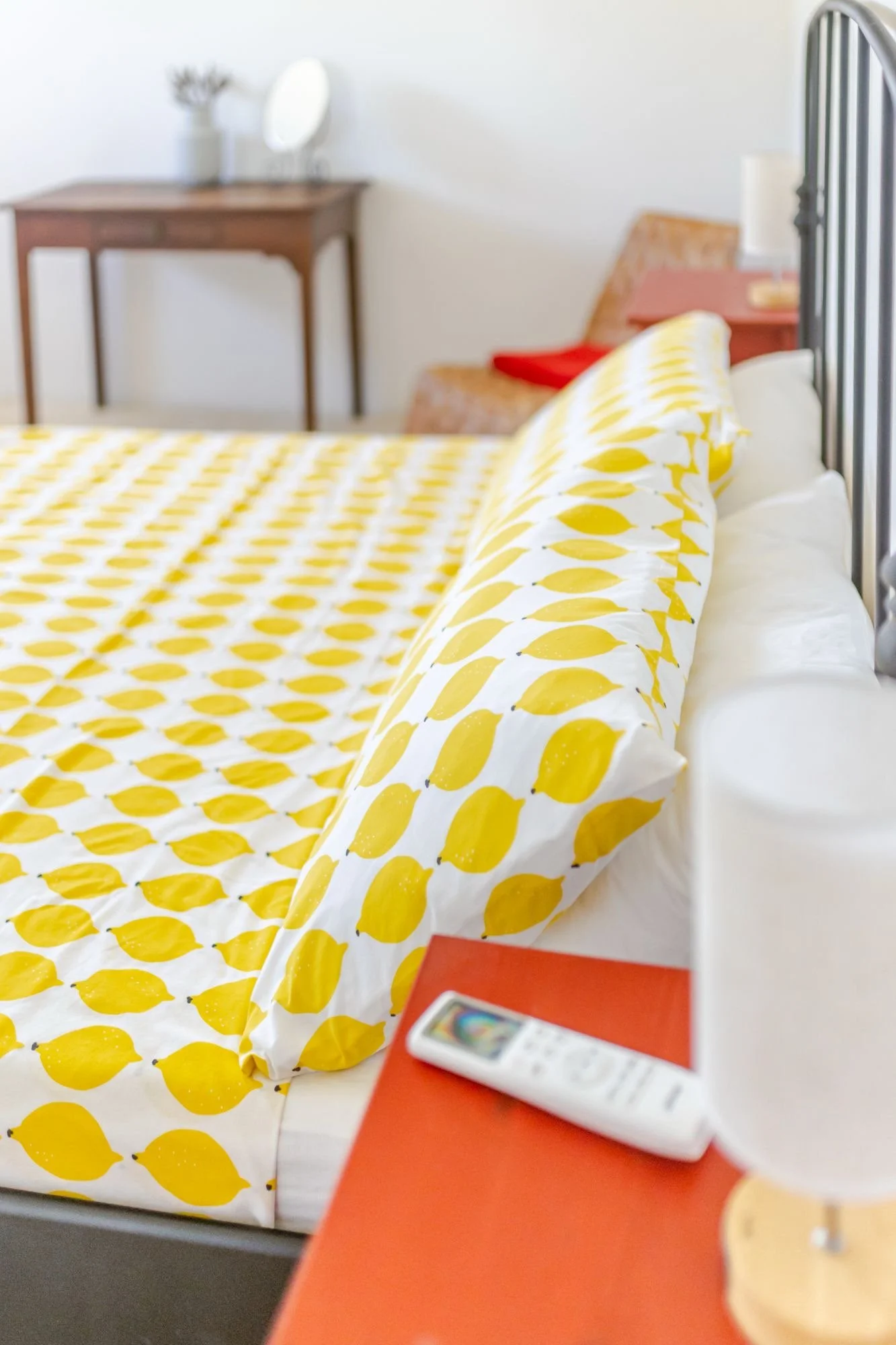 A bedroom with a bed covered in white and yellow patterned bedding. In the background, there is a wooden desk with a mirror and decorative items. A red book and a remote control are on a red surface at the foreground.