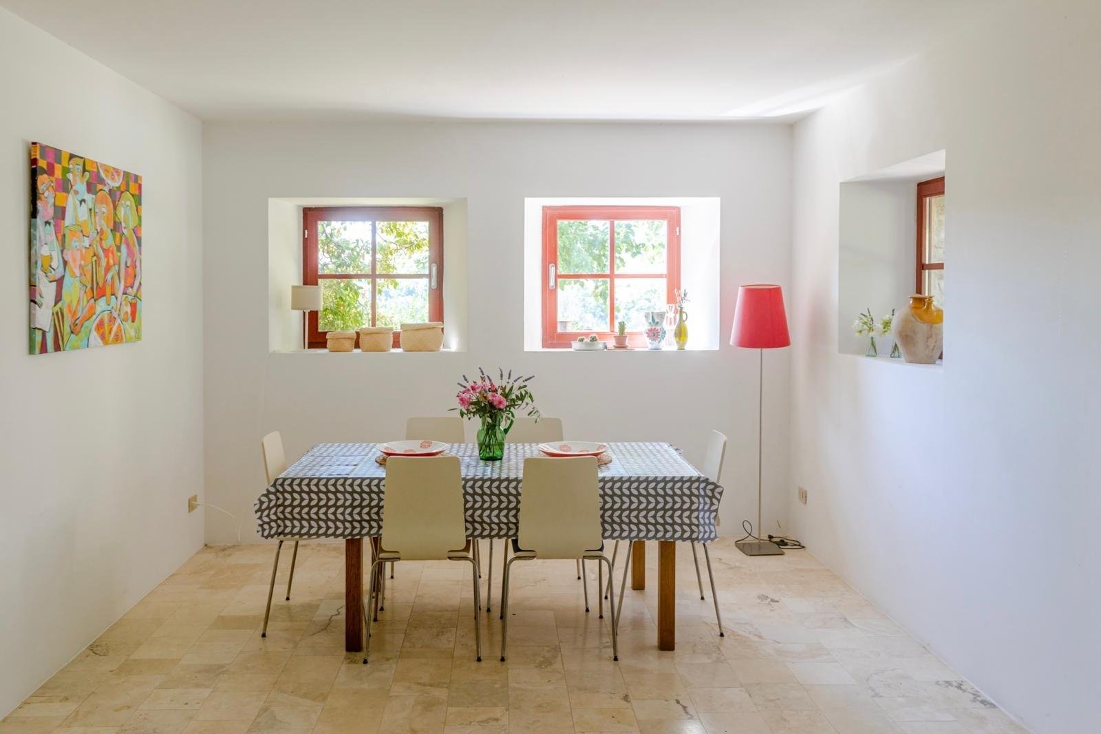 Bright dining area with white walls, three small windows with red frames letting in natural light, a table with a black and white patterned tablecloth, five white chairs, a vase with pink flowers, and decorative items on the windowsills, including a 