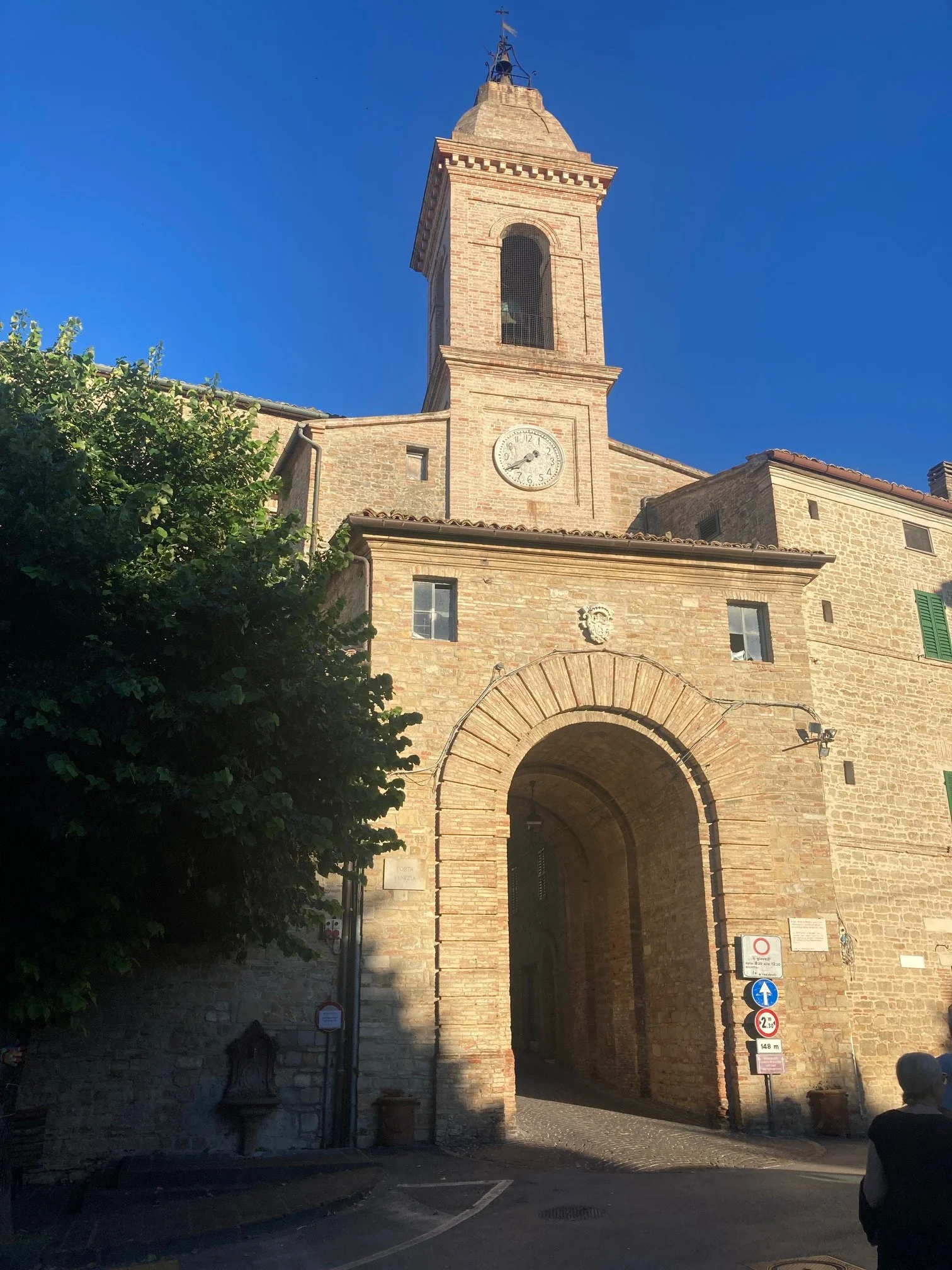 Bridge with a brick archway and tower, clock on the tower, signs below, and cobblestone street, with trees on the side and a person in the foreground.