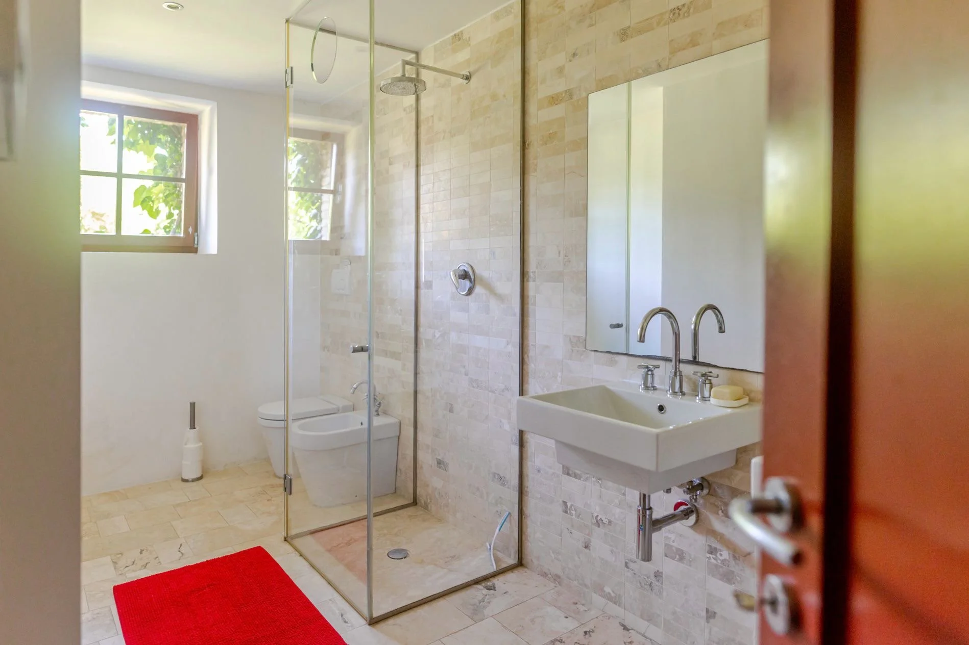 A modern bathroom with a glass-enclosed shower, a wall-mounted sink with a mirror above, two windows letting in natural light, a toilet with an automatic bidet, and a red rug on the tile floor.