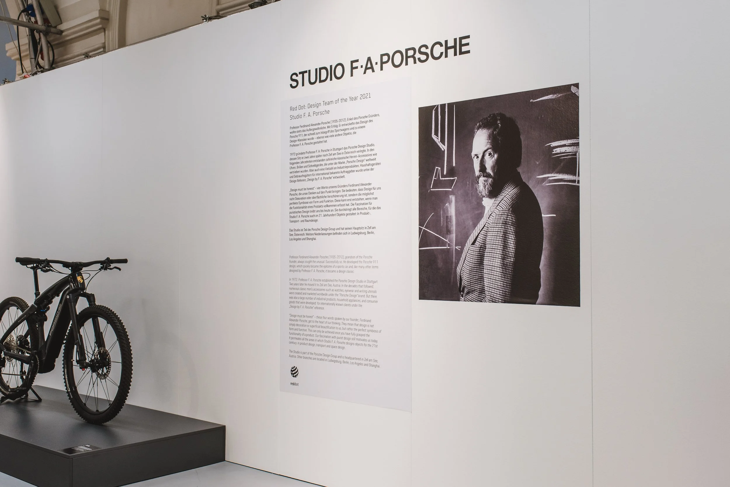 Ein Ausstellungswand mit Informationen über Studio F. A. Porsche, darunter ein schwarzes und weißes Porträt eines Mannes, und ein Fahrrad ist im Vordergrund sichtbar.