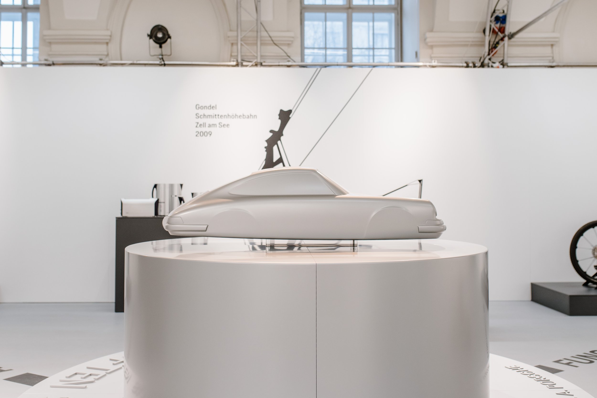 Ein weißes, futuristisches Fahrzeug auf einem weißen Podium in einem hellen Ausstellungsraum mit hohen Fenstern und weißer Wand, auf der eine Skizze eines Gondel-ähnlichen Fahrzeugs und der Text 'Gondel Schittenhoebahn Zell am See 2009' zu sehen sind.