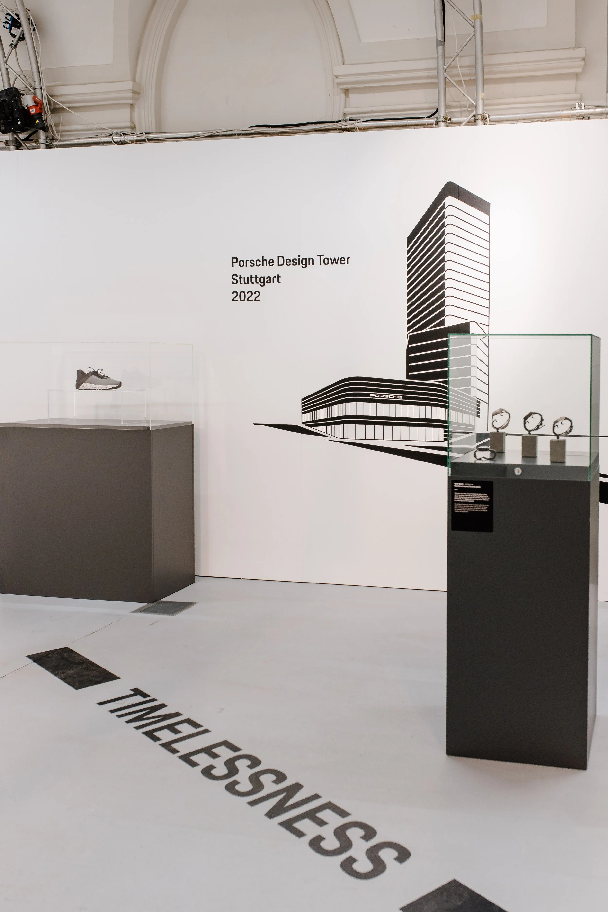 Ausstellung mit dem Schriftzug "Porsche Design Tower Stuttgart 2022", eine Skizze des Gebäudes, eine ausgestellte Sportschuh und Uhren auf schwarzen Ständern.