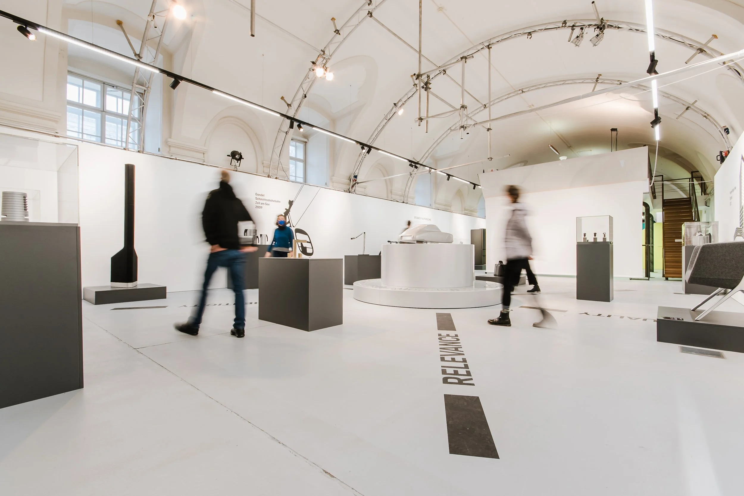 Innenansicht eines modernen Museums oder Ausstellungsraumes mit weißen Wänden und hohen Decken. Mehrere Besucher laufen durch den Raum, einige mit Masken. Auf Displays und Podesten sind Kunstwerke oder Objekte ausgestellt, und es gibt große Fenster, die natürliches Licht lassen. Die Raumgestaltung ist minimalistisch und zeitgenössisch.