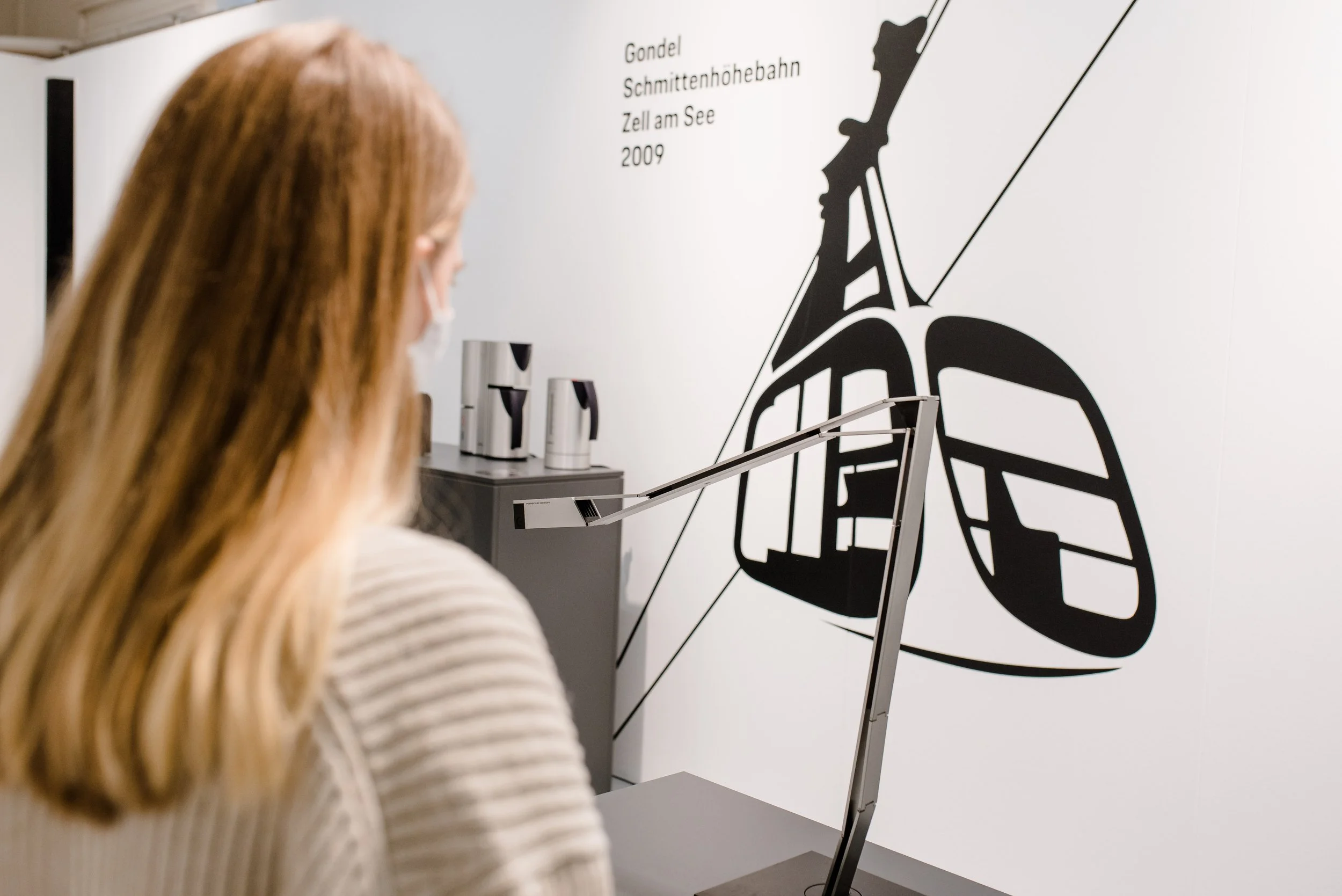Eine Person mit langen, roten Haaren betrachtet ein moderner, schwarzer und weißer Wand- und Stehlampen-Poster in einem Museum oder Ausstellung. Das Poster zeigt ein stilisiertes, schwarzes Sessel-Design und ist mit deutschen Texten über eine Seilbahn-Attraktion versehen, darunter die Wörter 'Gondel', 'Schmittenhöhebahn', 'Zell am See' und das Jahr 2009.