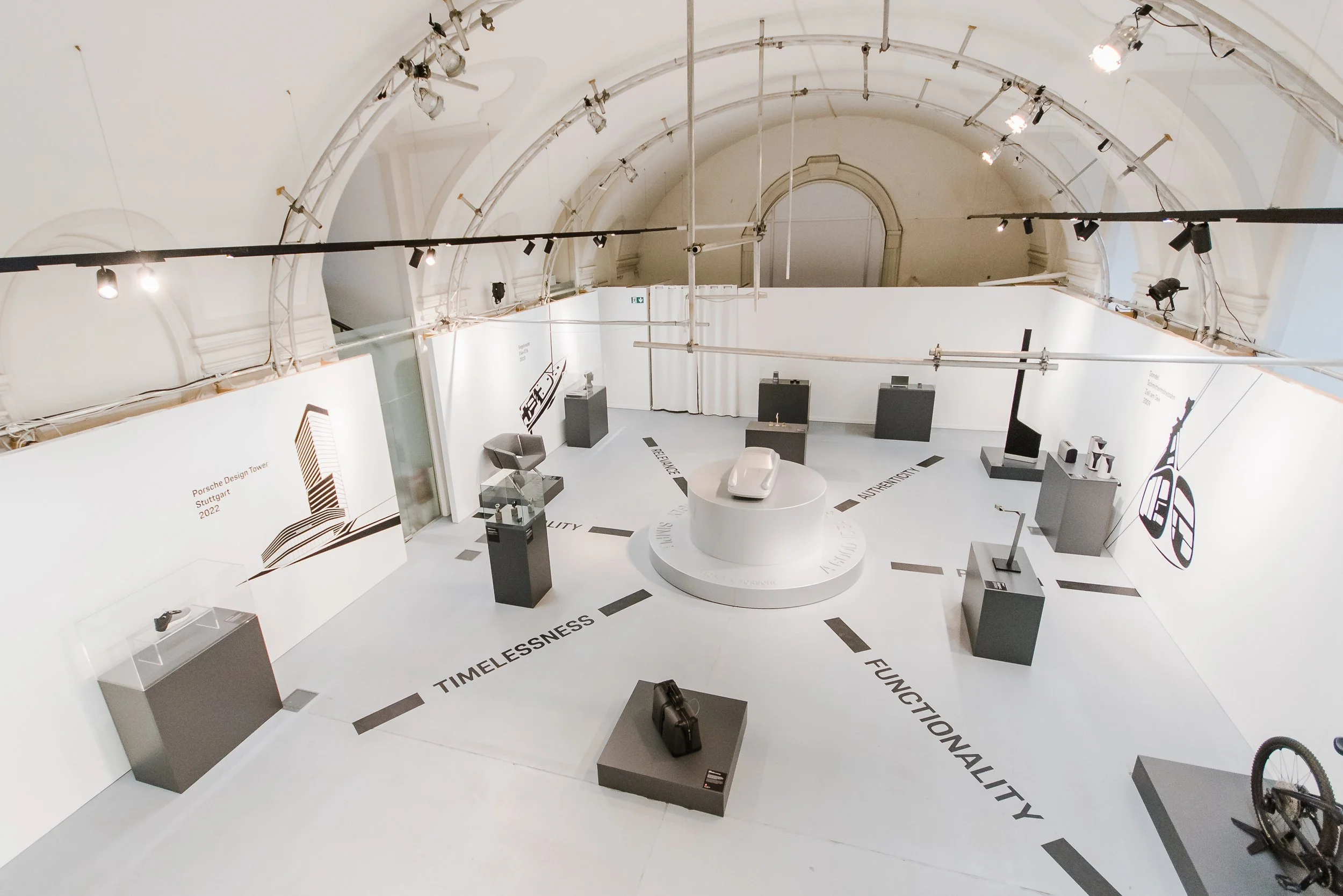 Ausstellung im Museum mit Automobilen und Design-Exponaten, weiße Wände und Lichtinstallationen, Bodenmarkierungen mit Begriffen wie "Timelessness", "Elegance", "Functionality".