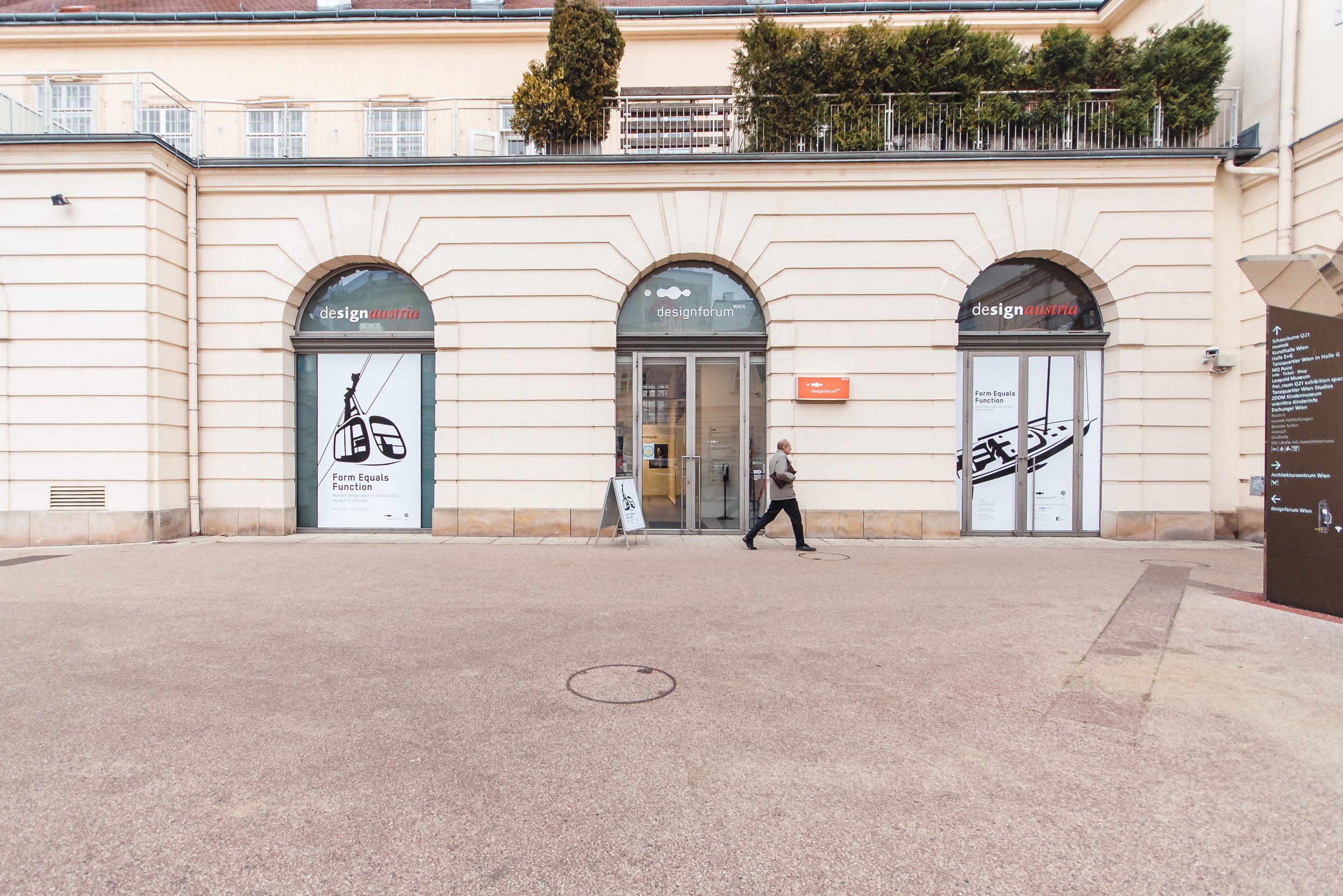 Außenansicht eines Gebäudes mit runden Fensteröffnungen, auf denen Anzeigen für eine Designmesse in Wien angebracht sind, sowie eine Person, die vorbeigeht.