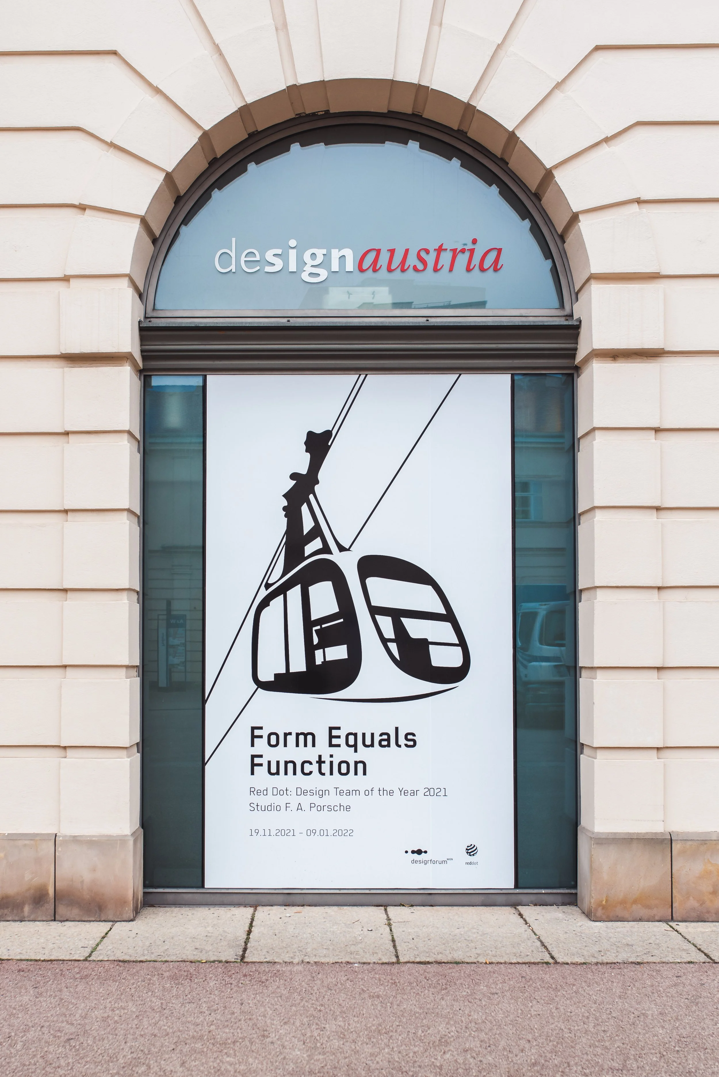 Plakat vor einem Gebäude mit dem Schriftzug "designaustria" im oberen Bereich und einer Illustration einer Seilbahn im unteren Bereich, das Text enthält: "Form Equals Function", sowie Angaben zu einer Ausstellung vom 19.11.2021 bis 09.01.2022, und Logos von designforum und red dot.